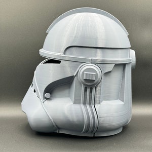 Puede incluir: Un casco gris impreso en 3D con un dise&ntilde;o detallado. El casco tiene una visera y una secci&oacute;n elevada en la parte superior.
