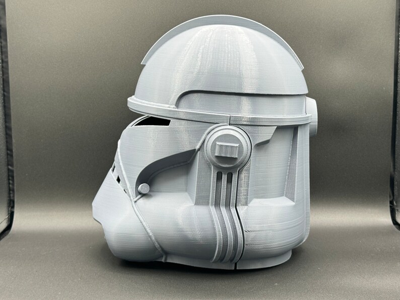 Puede incluir: Un casco gris impreso en 3D con un dise&ntilde;o detallado. El casco tiene una visera y una secci&oacute;n elevada en la parte superior.