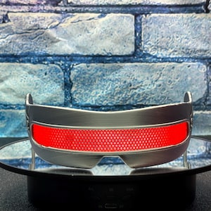 May include: Silver-colored, futuristic-style eyewear with a bright red, grid-patterned lens. The glasses have a curved design and are displayed on a black rotating stand. The background features a blue brick wall.