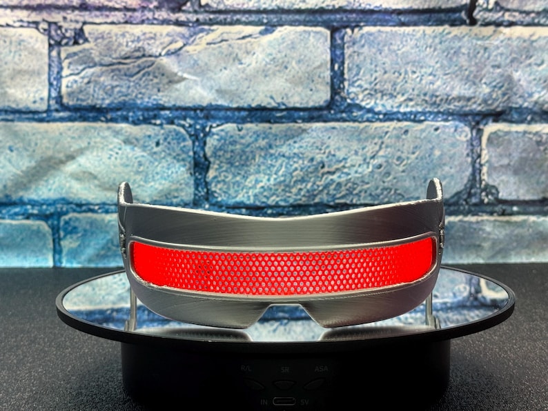 May include: Silver-colored, futuristic-style eyewear with a bright red, grid-patterned lens. The glasses have a curved design and are displayed on a black rotating stand. The background features a blue brick wall.
