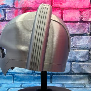 May include: A silver-colored helmet with a distinctive design, featuring a prominent vertical ridge and a curved visor. The helmet is displayed on a black stand against a brick wall with blue, pink, and purple hues.