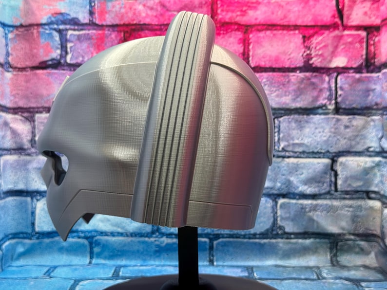 May include: A silver-colored helmet with a distinctive design, featuring a prominent vertical ridge and a curved visor. The helmet is displayed on a black stand against a brick wall with blue, pink, and purple hues.