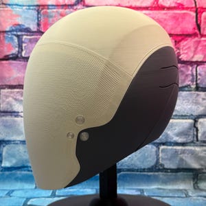 May include: A futuristic-style helmet in cream and dark gray, mounted on a black stand. The helmet has a textured surface on the cream-colored portion and smooth dark gray accents. Two silver-colored rivets are visible on the side.