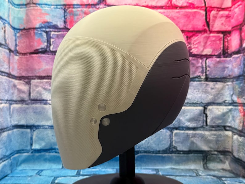 May include: A futuristic-style helmet in cream and dark gray, mounted on a black stand. The helmet has a textured surface on the cream-colored portion and smooth dark gray accents. Two silver-colored rivets are visible on the side.