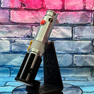 Pu&ograve; includere: Un'elica di spada laser argentata con dettagli neri e un pulsante rosso, esposta su un supporto nero. L'accessorio presenta un design dettagliato con elementi gialli e neri. Lo sfondo &egrave; un muro di mattoni con tonalit&agrave; rosa, blu e viola.