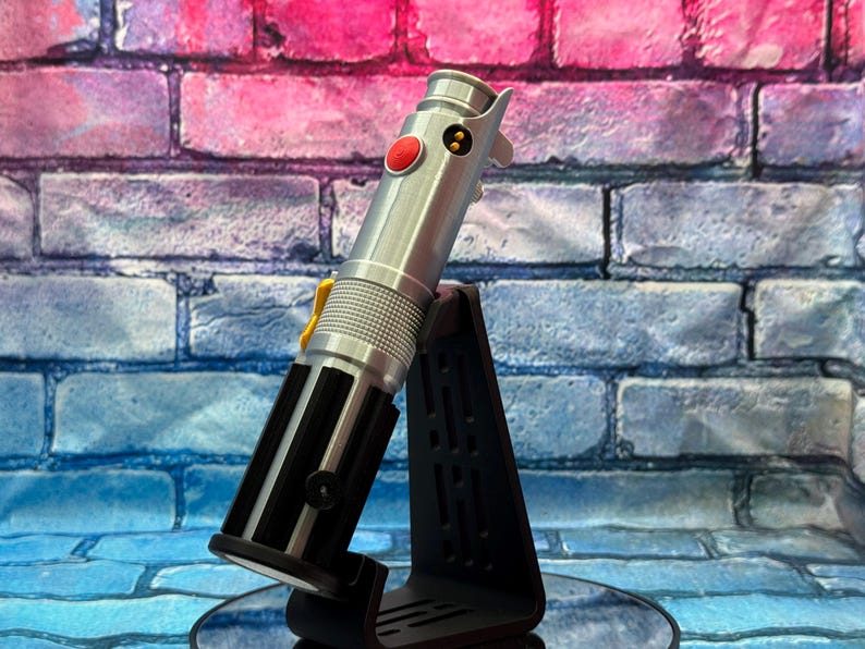 Pu&ograve; includere: Un'elica di spada laser argentata con dettagli neri e un pulsante rosso, esposta su un supporto nero. L'accessorio presenta un design dettagliato con elementi gialli e neri. Lo sfondo &egrave; un muro di mattoni con tonalit&agrave; rosa, blu e viola.