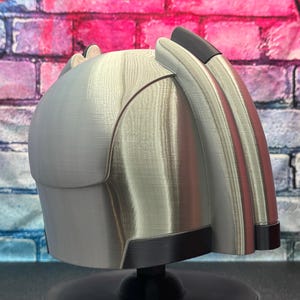 May include: A silver-colored helmet with black and red accents. The helmet has a curved design with a smooth, metallic finish. The helmet is displayed on a black stand against a brick wall.