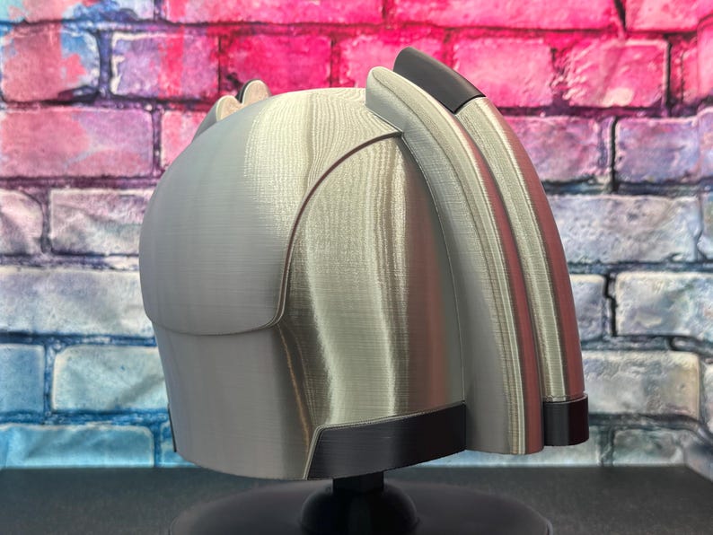 May include: A silver-colored helmet with black and red accents. The helmet has a curved design with a smooth, metallic finish. The helmet is displayed on a black stand against a brick wall.