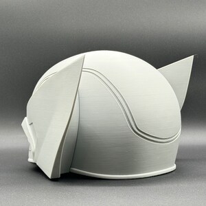 Puede incluir: Un casco blanco impreso en 3D con una parte superior redondeada y dos orejas puntiagudas a los lados. El casco tiene una superficie lisa y texturizada.