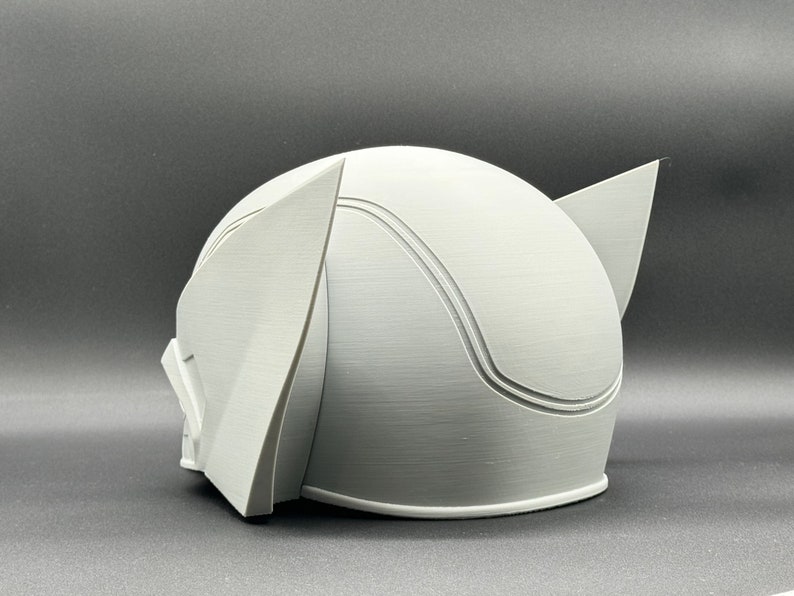 Puede incluir: Un casco blanco impreso en 3D con una parte superior redondeada y dos orejas puntiagudas a los lados. El casco tiene una superficie lisa y texturizada.