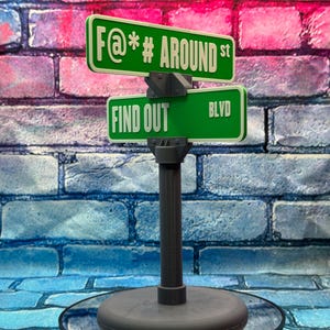 May include: A novelty street sign with two green signs. The top sign reads "F@*# AROUND st" and the bottom sign reads "FIND OUT BLVD". The sign is mounted on a gray post with a round base. The background is a brick wall.