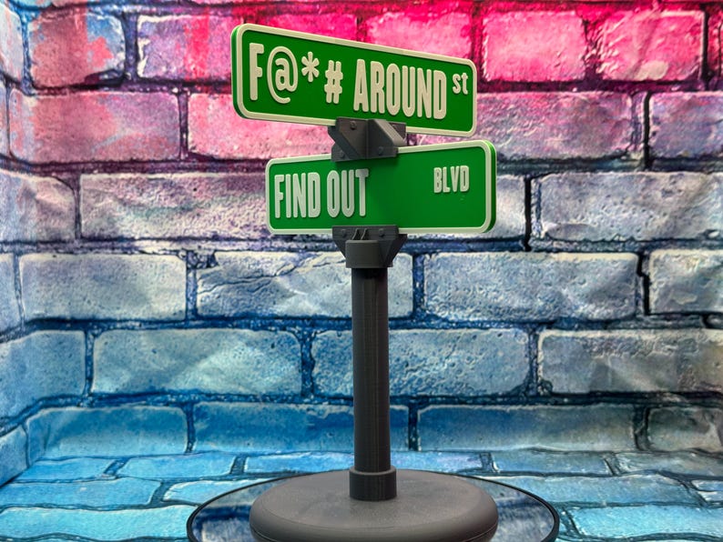 May include: A novelty street sign with two green signs. The top sign reads "F@*# AROUND st" and the bottom sign reads "FIND OUT BLVD". The sign is mounted on a gray post with a round base. The background is a brick wall.