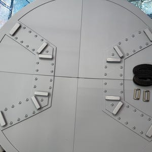May include: A large, circular, gray metal object with a segmented design. It features rectangular and triangular accents, rivets, and a black strap with two silver buckles. The object is likely a component of a larger structure.