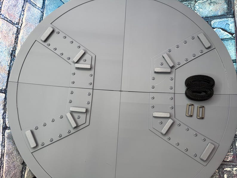 May include: A large, circular, gray metal object with a segmented design. It features rectangular and triangular accents, rivets, and a black strap with two silver buckles. The object is likely a component of a larger structure.