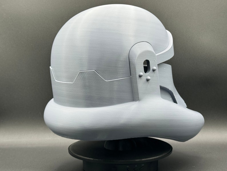 May include: A white plastic helmet with a textured surface. The helmet has a rounded top and a curved bottom. The helmet has a small hole on the side and a series of small bumps on the back.