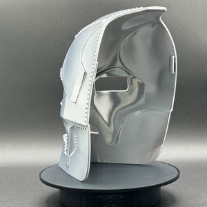 May include: A white 3D printed mask with a textured surface. The mask is open on one side and has a small hole on the other side. The mask is sitting on a black circular base.