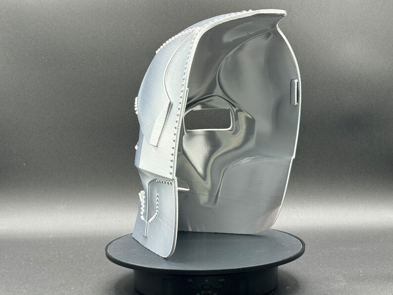 May include: A white 3D printed mask with a textured surface. The mask is open on one side and has a small hole on the other side. The mask is sitting on a black circular base.