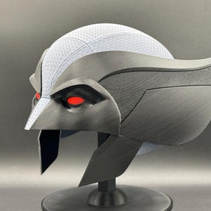 May include: A black and gray helmet with red eyes. The helmet has a textured surface and a wing-like design on the side.