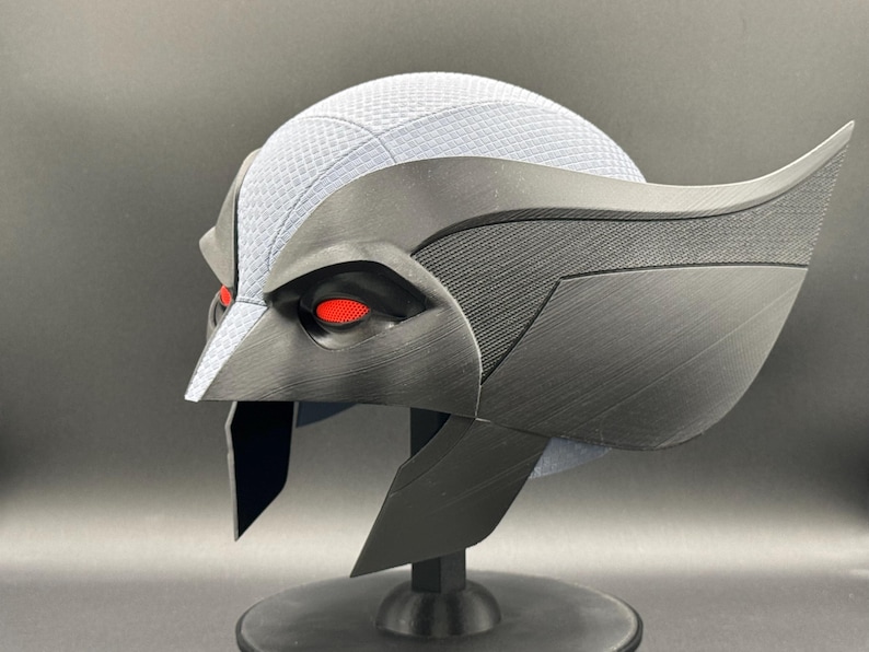 May include: A black and gray helmet with red eyes. The helmet has a textured surface and a wing-like design on the side.