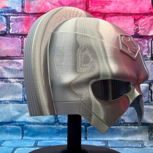 May include: A silver and red helmet with a stylized design, featuring a prominent eye opening and a crest emblem. The helmet is displayed on a black stand against a brick wall background.