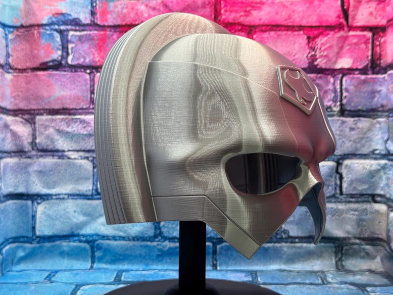 May include: A silver and red helmet with a stylized design, featuring a prominent eye opening and a crest emblem. The helmet is displayed on a black stand against a brick wall background.
