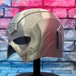 May include: A silver and red helmet with a distinctive design, possibly for cosplay or display. The helmet features a stylized shape with a prominent visor area and a crest detail. It is mounted on a black stand against a brick wall background.