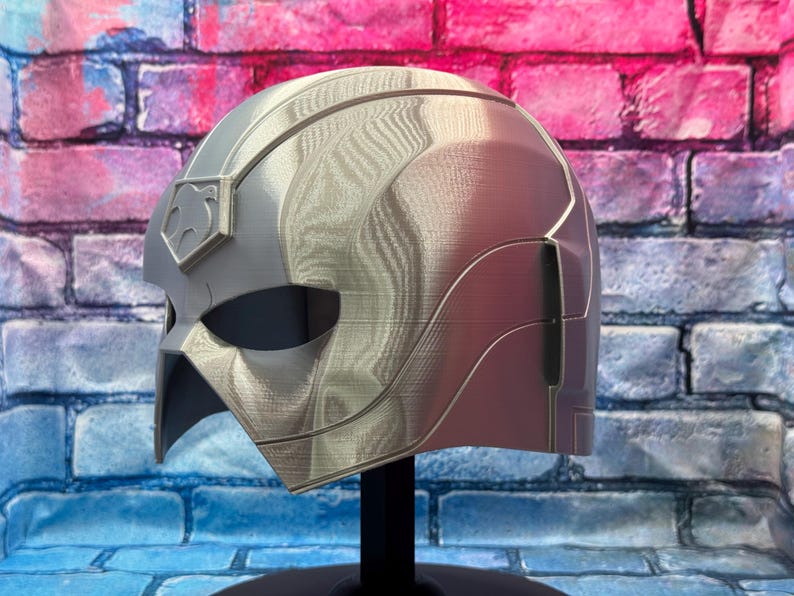 May include: A silver and red helmet with a distinctive design, possibly for cosplay or display. The helmet features a stylized shape with a prominent visor area and a crest detail. It is mounted on a black stand against a brick wall background.