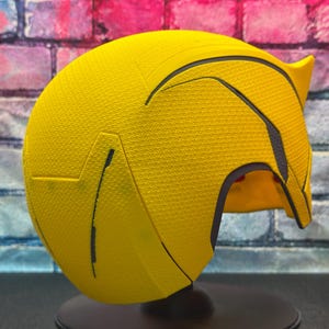 Puede incluir: Un casco amarillo con detalles negros y superficie texturizada. El casco tiene protuberancias en forma de oreja y una visera curva. El interior es rojo. El casco se exhibe sobre un soporte negro. Ideal para cosplay.