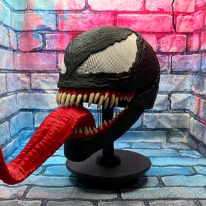 May include: A black Venom mask with a white mesh eye area, sharp teeth, and a long, red tongue. The mask is displayed on a black stand. The mask is a replica of the Marvel Comics character.