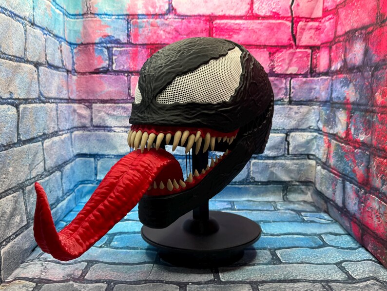 May include: A black Venom mask with a white mesh eye area, sharp teeth, and a long, red tongue. The mask is displayed on a black stand. The mask is a replica of the Marvel Comics character.