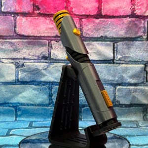 May include: A gray and yellow lightsaber prop, with a black stand, against a brick wall backdrop. The lightsaber features a ribbed yellow emitter and yellow buttons. The stand is black and angled.