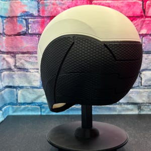 May include: A two-tone helmet, with a white upper section and a black lower section. The black section has a textured, geometric pattern. The helmet is displayed on a black stand against a colorful brick wall background.