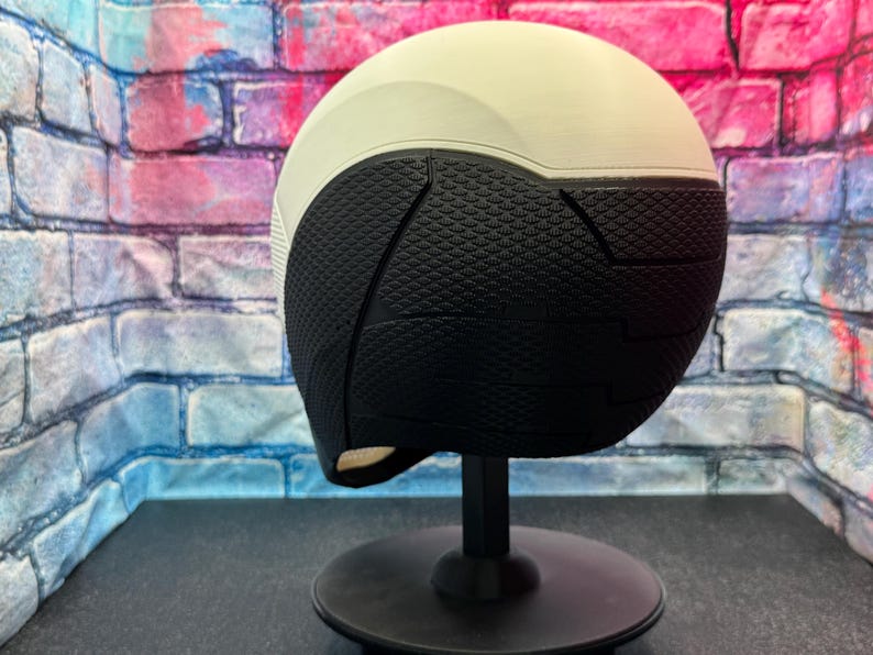 May include: A two-tone helmet, with a white upper section and a black lower section. The black section has a textured, geometric pattern. The helmet is displayed on a black stand against a colorful brick wall background.