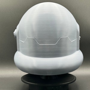 May include: A white 3D printed helmet with a rounded top and a textured surface. The helmet has a raised design around the bottom edge.