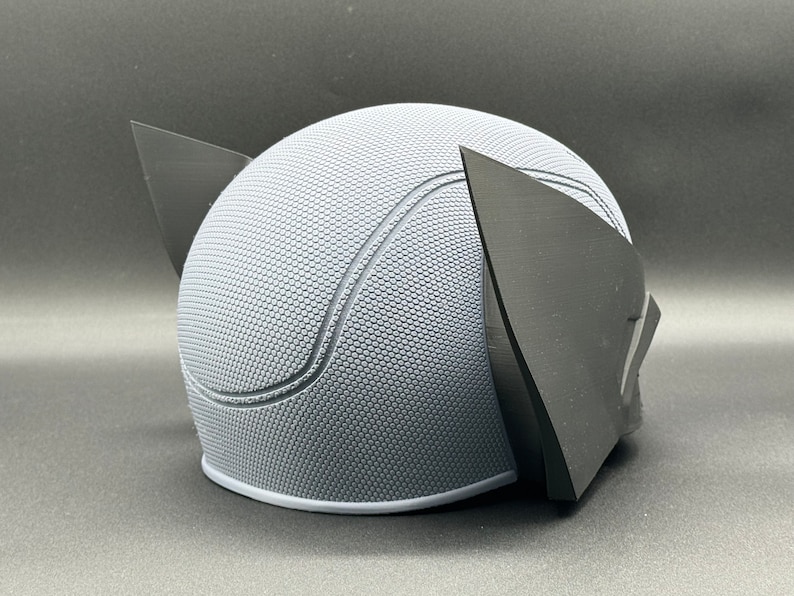 May include: A gray and black 3D printed helmet with a textured surface and two pointed ears.