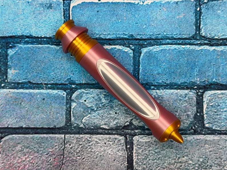 Op de afbeelding: Een roze en goudkleurig penvormig object met een zilverkleurig ovaal ontwerp. De pen heeft een puntige punt en een geribbeld gouden gedeelte aan de bovenkant. De achtergrond is een blauw en grijs baksteenpatroon.