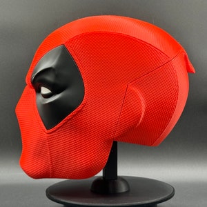 Puede incluir: Una r&eacute;plica de m&aacute;scara de Deadpool roja y negra. La m&aacute;scara est&aacute; en un soporte negro y se ve de lado. La m&aacute;scara tiene una superficie texturizada y una m&aacute;scara para los ojos negra.