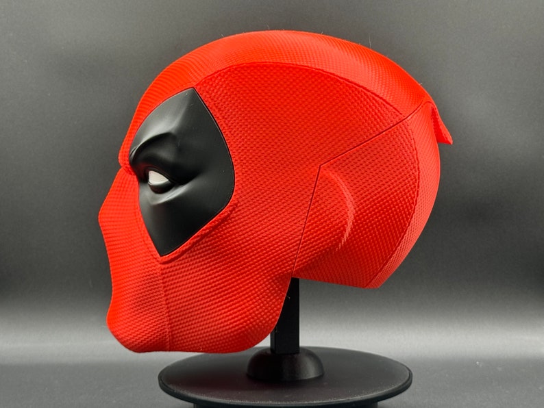 Puede incluir: Una r&eacute;plica de m&aacute;scara de Deadpool roja y negra. La m&aacute;scara est&aacute; en un soporte negro y se ve de lado. La m&aacute;scara tiene una superficie texturizada y una m&aacute;scara para los ojos negra.