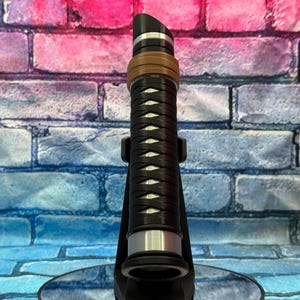May include: A black and silver lightsaber prop with a brown and silver accent ring. The hilt features a woven design. The lightsaber is displayed on a black stand against a colorful brick wall background.