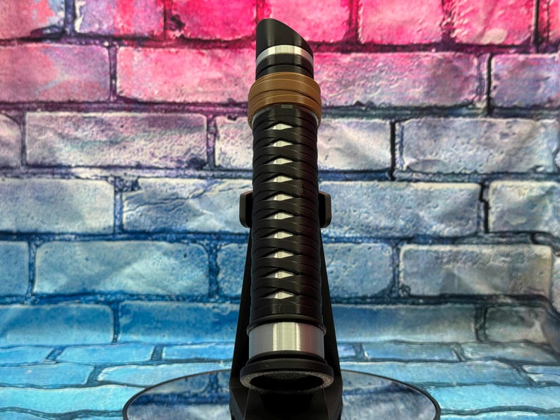 May include: A black and silver lightsaber prop with a brown and silver accent ring. The hilt features a woven design. The lightsaber is displayed on a black stand against a colorful brick wall background.