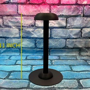 May include: Black hat display stand, 11 inches tall, with a round base and a curved top. The stand is designed to hold and showcase hats. The background is a brick wall with blue, pink, and purple hues.