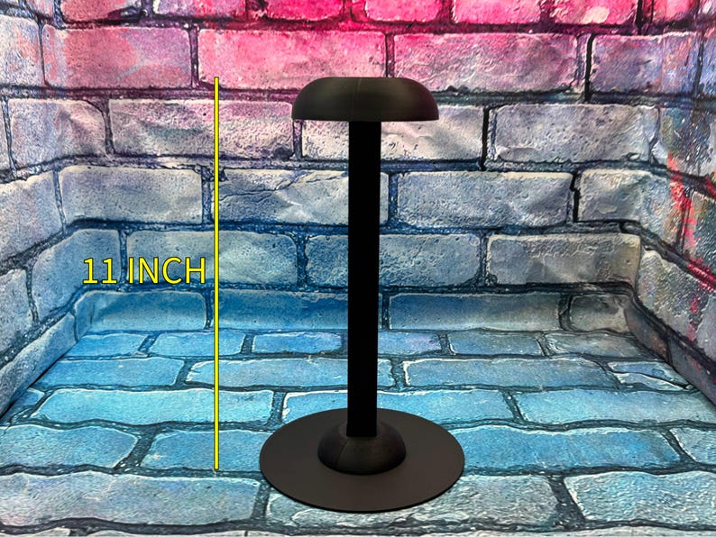 May include: Black hat display stand, 11 inches tall, with a round base and a curved top. The stand is designed to hold and showcase hats. The background is a brick wall with blue, pink, and purple hues.