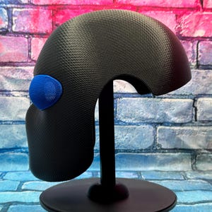 May include: A black helmet-like headpiece with a textured carbon fiber pattern and a single bright blue oval accent on the side. The object is mounted on a black vertical stand with a circular base, displaying a smooth, curved, and aerodynamic shape.