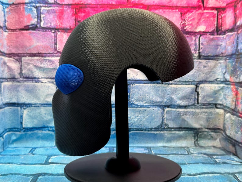 May include: A black helmet-like headpiece with a textured carbon fiber pattern and a single bright blue oval accent on the side. The object is mounted on a black vertical stand with a circular base, displaying a smooth, curved, and aerodynamic shape.