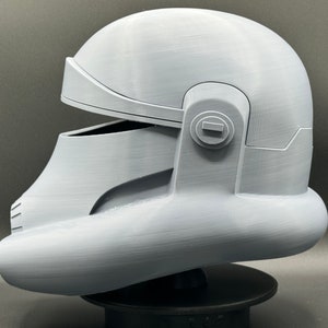 May include: A white 3D printed helmet with a visor and a round detail on the side. The helmet is on a black stand.