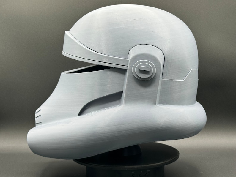 May include: A white 3D printed helmet with a visor and a round detail on the side. The helmet is on a black stand.