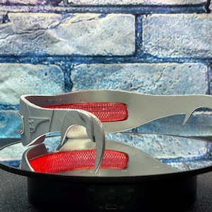 May include: A silver-colored, futuristic-style headpiece with a red mesh insert. The headpiece has a curved design with pointed ends and is displayed on a reflective surface. The background is a blue brick wall.