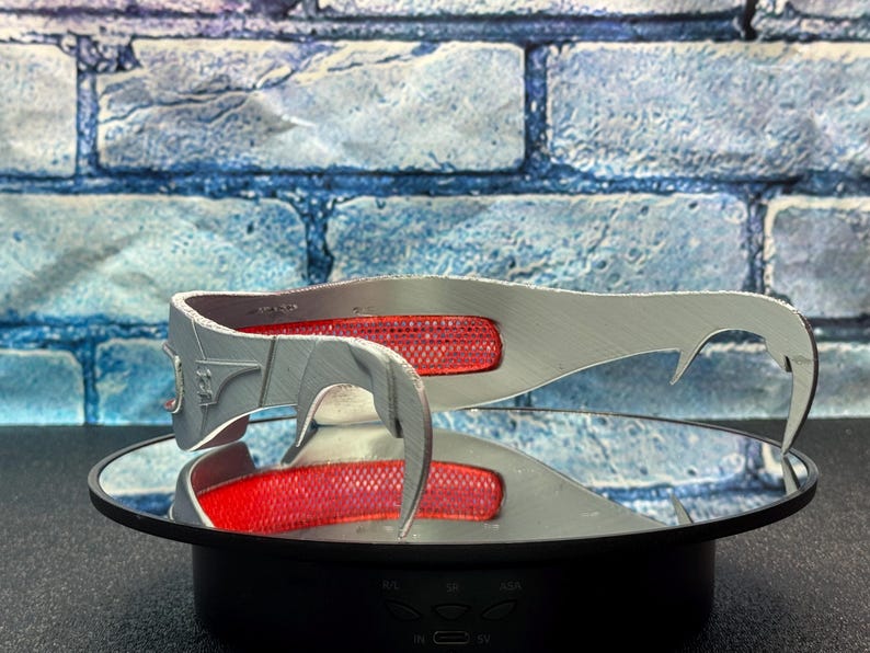 May include: A silver-colored, futuristic-style headpiece with a red mesh insert. The headpiece has a curved design with pointed ends and is displayed on a reflective surface. The background is a blue brick wall.