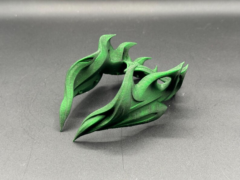 Puede incluir: Dos objetos verdes abstractos impresos en 3D con un dise&ntilde;o org&aacute;nico y fluido. Los objetos tienen una serie de elementos curvos y puntiagudos, y est&aacute;n colocados sobre una superficie gris. Los objetos parecen decorativos.