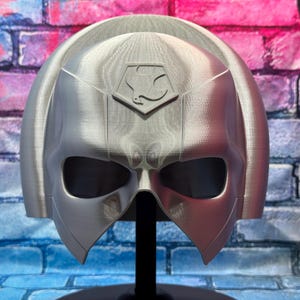 May include: A silver-colored helmet with a distinctive design, featuring a prominent eye-shaped opening and a crest with a stylized emblem. The helmet is displayed on a black stand against a brick wall background.